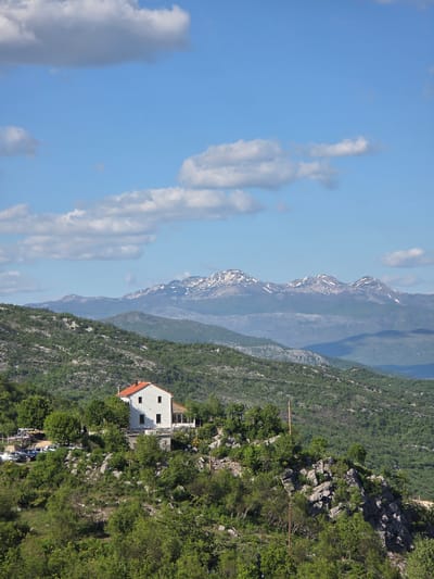 The image shows a mountainous landscape in Ponikvica, Montenegro, with a white building featuring a red roof situated on a hillside covered with lush green vegetation. In the background, a mountain range is visible, with some snow-capped peaks. The sky is blue with scattered white clouds. There are parked cars near the building.