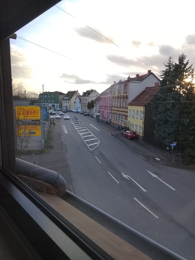 The image presents an elevated view of a street in Neunkirchen, Germany, observed through a window frame with a visible sill and pipe in the foreground. The scene is illuminated by the low sun, visible on the right, under a partly cloudy sky, suggesting late afternoon or early evening. A two-lane asphalt road occupies the mid-ground, featuring white dashed and solid lane lines, a hatched white median strip, and white directional arrows indicating straight and left-turn options.

On the left side of the street, a large yellow "Netto Marken-Discount" billboard is prominently displayed, behind which several vehicles are parked. Further down the street on the left, multi-story buildings with varied facades (green, beige) are visible. On the right side of the street, a row of multi-story buildings features facades in pink, beige, and yellow, all with reddish-brown tiled roofs. A large conifer tree on the far right partially obscures the sun. A red car is parked on the right side of the road near a blue directional sign. Several other vehicles, including at least two white cars, are present on the road further down the street. No individuals are distinctly visible or identifiable.