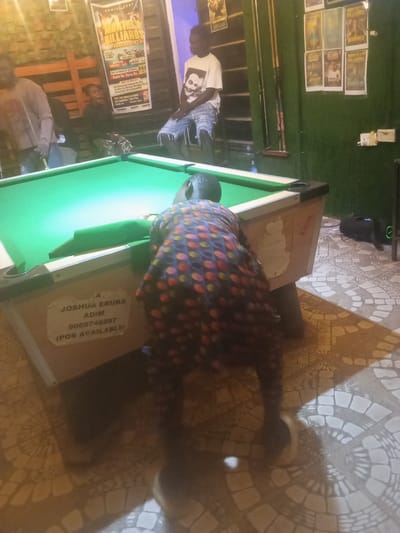 The image depicts an indoor scene at a pool hall in Ijegun, Nigeria. In the foreground, a person in a colorful patterned shirt is bent over a pool table, appearing to be playing. The pool table is green with black and white accents. To the left of the table, a sign is visible with text that includes "JOSHUA EBUKA," "ADIM," a phone number, and "(POS AVAILABLE)." In the background, several individuals are present, including one sitting on a stool wearing a white t-shirt with a skull graphic and distressed denim shorts, and another standing on the left wearing a grey long-sleeved shirt. The walls are decorated with posters, some advertising "MONTHLY BILLIARDS." The flooring is tiled with a patterned design. The overall lighting suggests an evening or indoor setting.