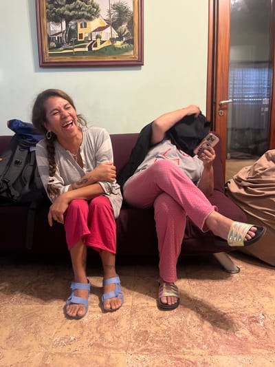 Two people are sitting on a dark maroon-colored couch. One person is looking at a smartphone and is wearing pink polka dot pajamas, a grey shirt, and Adidas-branded black sandals with iridescent stripes. The second person is sitting on the other end of the couch, laughing. She is wearing a grey and white striped shirt, pink shorts, and blue sandals. A grey and blue backpack is sitting near her. A framed painting of a house is on the wall behind her. A light brown window is in the background behind the first person. The floor is marbled in shades of light brown and tan. There is no information to suggest that this photograph was taken in Saladillo, Argentina.