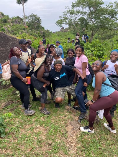 A group of approximately 15-20 people are gathered outdoors. They are positioned in close proximity, with some individuals lying on top of others. The group appears to be a mix of genders and age ranges.
People are wearing a variety of clothing, including t-shirts, leggings, jeans, athletic wear, and head coverings. Some are holding items such as water bottles, hats, and a mobile phone.
The setting appears to be a grassy area with trees and rocks visible in the background. The image was taken in the city of Zankan, Nigeria.