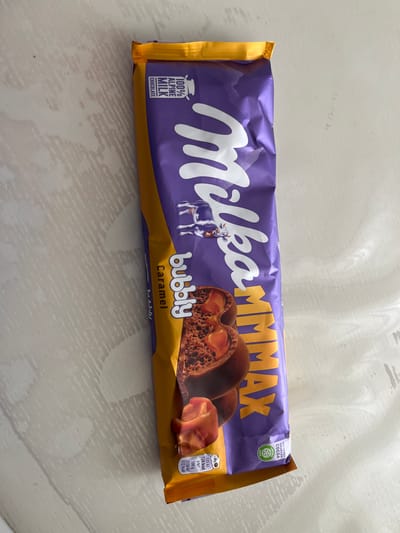 The image shows a Milka brand chocolate bar lying on a white, textured surface. The chocolate bar has purple packaging with a yellow border. The Milka logo, featuring a cow, is prominently displayed on the front. Text on the packaging includes "100% ALPINE MILK CHOCOLATE", "bubbly", "Caramel", and "MAX". There is an image of the chocolate bar itself, showing its bubbly texture and caramel filling. A small circular icon with text is also visible near the bottom right corner. The lighting suggests an indoor environment. The location is not discernable from the image.