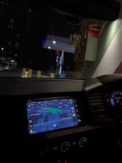 The image shows the dashboard of a car at night. The digital display shows a navigation map indicating the location as Sandton, Johannesburg, South Africa. Specifically, the route includes Rivonia Road (M9), Katherine Street (M85 E), and Daisy Street. The car is in park (P), with 9.7 km travelled. The A/C controls are visible, with some knobs lit in blue and red. The rear-view mirror and an obstructed view through the windshield are also present. The time is 0:11.