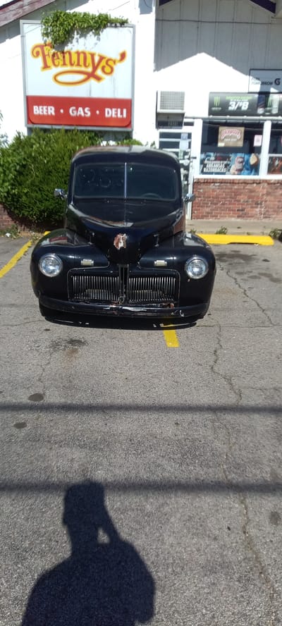 The image shows a black vintage car parked in front of a "Tennys" store in Fort Smith, Arkansas. The sign above the store advertises "Beer Gas Deli." The car is facing the camera and is parked on a yellow parking space line. The pavement is gray and cracked with dark oil stains. The shadow of the person taking the photo is visible in the foreground. The store has signage and other commercial displays visible through its windows.