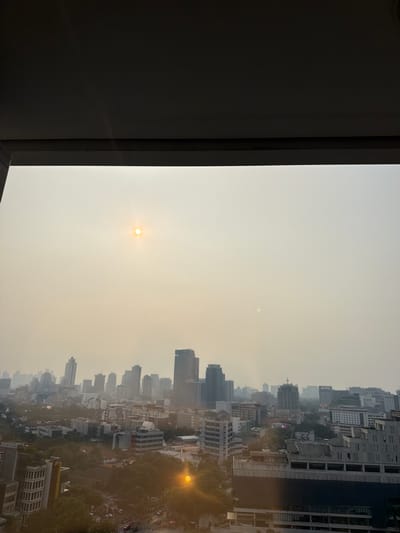 The image depicts a hazy skyline of Jakarta, Indonesia, as viewed from a high vantage point, possibly a balcony or window, framed by a dark overhang. The sun, a bright orb, is visible in the upper left quadrant, partially obscured by atmospheric haze, giving it a soft glow. The haze permeates the entire scene, softening the edges of buildings and creating a muted color palette.
Below the hazy sky, a dense urban landscape unfolds. Numerous buildings of varying heights and architectural styles dominate the view. There are prominent skyscrapers with modern designs, interspersed with lower-rise buildings. The architecture suggests a developed city center. Greenery, in the form of trees and possibly parks, can be seen peeking through the urban structures, particularly in the mid-ground.
The foreground shows the edge of a building or structure, contributing to the framing of the cityscape. The overall impression is of a busy city, where the atmospheric conditions, characterized by the haze, significantly impact visibility and the quality of light. The lower part of the image shows hints of street level activity, with what appear to be vehicles and potentially some pedestrian presence, though they are indistinct due to the distance and haze. The presence of the sun and the general brightness of the sky suggest it is daytime, likely late morning or early afternoon.