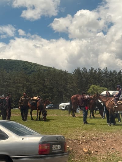 The image shows a scene near Gyulagarak, Armenia. Several individuals on horseback are present. A wooded hillside forms the backdrop. A silver car is parked nearby, its license plate partially visible. The sky is partly cloudy. The ground is covered in grass and some bare earth.
