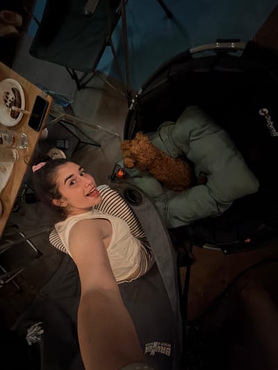 The image depicts a dimly lit indoor scene, possibly a campsite or a room with camping equipment. A young woman with dark hair tied up and a pink scrunchie is lying down, looking at the camera and sticking her tongue out. She is wearing a white tank top with black stripes and dark pants. To her right, a small brown dog, possibly a poodle mix, is nestled in a folded green sleeping bag or blanket. The background includes camping chairs and a black Outwell branded item, likely a tent or a chair, with a grey handle. On a wooden surface to the left, there is a plate with food, a mobile phone, and some glasses. The overall impression is a casual, relaxed moment captured at night. There is no indication of the location being in Πόρτες, Greece.