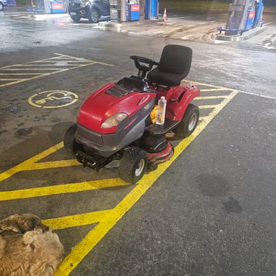 A red and gray riding lawn mower sits in a parking space marked with yellow lines and a disabled access symbol at a gas station, with a car parked at a pump and a brown dog in the lower right corner of the image. The location appears to be the city of Sopot Urban Municipality, Serbia.