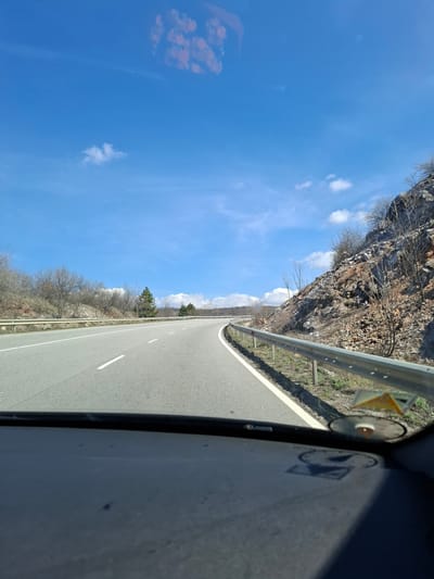 An eye-level, canted, mobile phone photograph, from inside a vehicle, shows an empty, two-lane asphalt road in Bogyovtsi, Bulgaria. To the left of the road is a bright, blue sky with a few small, white clouds, and the road is flanked by an aging guardrail. On the left side of the road, beyond the guardrail, is a rocky hillside. On the right side of the road, beyond the guardrail, is a dry, forested area. In the lower right of the photograph is the inside door frame of the vehicle. The scene is during the day.