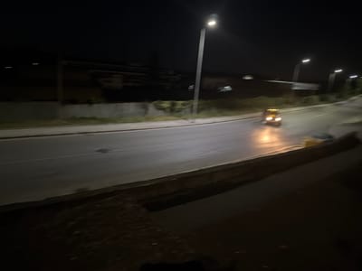 This image depicts a nighttime scene on a road in Peshtera, Bulgaria. A car with its headlights illuminated is driving on the asphalt road. Streetlights are visible in the background, casting light on the surroundings. Buildings and vegetation are also present along the roadside. The overall environment suggests a quiet urban or suburban area at night.