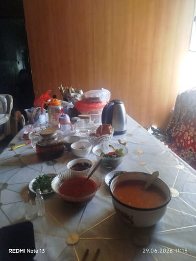 A table laden with food is set against a backdrop of wood-paneled walls. On the table, various dishes are arranged, including bowls of soup, a salad in a glass bowl, chopped herbs on a plate, and jars of what appears to be preserves or sauces. There are also cookies in a glass dish and a modern electric kettle. To the left, a child's high chair is partially visible. The scene suggests a domestic setting, possibly a home in Tokmok City, Kyrgyzstan, with a meal in progress or recently concluded.