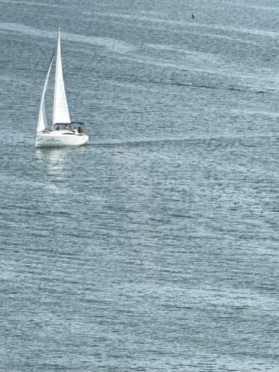 This is a high-angle, full shot of a small white sailboat with two people on board, navigating a body of water. The water appears to be a calm, grey surface, with a texture that resembles falling water or a dense mist, giving the impression of a waterfall or a very still, slightly obscured body of water. The sailboat has a prominent white sail, tilted to the left of the frame, and a dark-colored hull. The two individuals are visible in the cockpit of the boat. The overall scene is serene, with the soft, uniform color palette dominated by shades of grey and white. There are no discernible location cues within the image itself, other than what is provided in the context that the location is Mikołajki, Poland. No text is visible in the image. The time of day and weather are not explicitly indicated, but the diffused light suggests it could be overcast or early morning/late evening. The image has a painterly quality, with visible brushstroke-like textures, especially in the background water.