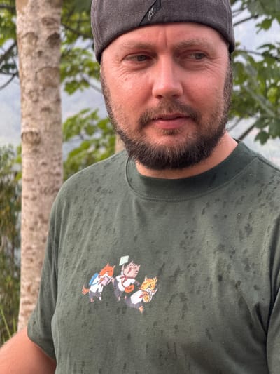 A man wearing a gray baseball cap and a green t-shirt with an illustration of three cat characters, possibly cartoon characters, stands outdoors. The t-shirt has visible spots or stains. The man has a beard and appears to be looking at the camera. Behind the man is a view of trees and a landscape with a hint of water, likely in a natural environment. The location is identified as Hoàng Su Phì Commune, Vietnam. The image is oriented sideways.