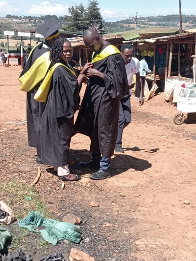 The scene depicts an unpaved outdoor area in Waseges ward, Kenya, under clear daylight conditions. In the center, a woman with braided hair, wearing a black academic gown and a yellow stole, faces a man with a shaved head, who is similarly attired in a black academic gown and yellow stole. The woman is actively adjusting an item on the man's chest area, while he looks downwards at the point of interaction. To the immediate left, an individual is partially visible from the back, also wearing a black academic cap, gown, and yellow stole. Behind the central pair, a fourth man in a white t-shirt and dark pants stands, observing the interaction.
The background features a row of informal market stalls constructed from wood and corrugated iron, some displaying hanging bananas. To the right, a small, white two-wheeled vendor's cart is visible, partially labeled "NICE B" and "NICE M." The ground is predominantly reddish-brown earth mixed with gravel, featuring scattered rocks and patches of green vegetation. In the lower left foreground, a crumpled light green fabric or plastic material and dark remnants consistent with charcoal are present. In the distant background, a hilly landscape with sparse trees and scattered structures is visible under a light blue sky.