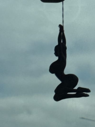A silhouette of a person is hanging from a thin cord against a cloudy sky. The figure is in a kneeling position with their arms raised above their head. The figure appears to be a statue or a decorative object, possibly hanging from a building or tree. The location in Didino, Russia is not discernible from the image.