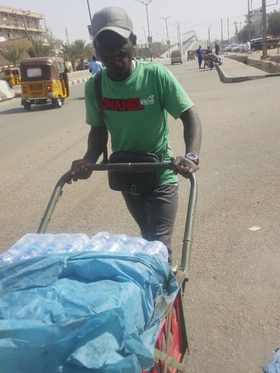 A dark-skinned male is depicted pushing a two-wheeled utility cart along a paved road in Gyadi Gyadi Arewa, Nigeria, under a hazy daylight sky. He wears a grey baseball cap featuring a Nike logo, a green short-sleeved t-shirt with "CHANEL" in red lettering and an "AFSCME" logo, dark trousers, and a black crossbody bag across his chest. A watch is visible on his left wrist, and red polish is present on the fingernails of his right hand as he grips the cart's metal handle. His mouth is slightly open. The cart, constructed with a red and green metal frame, carries multiple clear plastic bundles of bottled water, partially obscured by a light blue tarp.
The background reveals a bustling urban street. A yellow three-wheeled auto-rickshaw is visible directly behind the man on the left side of the road, with other vehicles, including additional auto-rickshaws, cars, and motorcycles, further in the distance. Concrete road dividers separate traffic lanes. Streetlights with multiple arms and a prominent elevated road or overpass structure are visible in the mid-ground. Multi-story buildings line both sides of the distant road, interspersed with palm trees, indicative of an urban setting. The road surface appears dry and dusty, with unpaved shoulders and exposed concrete drainage on the right.