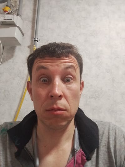 A close-up, head-and-shoulders selfie of a man with dark, short hair and light skin is presented. He has wide, surprised-looking eyes and his mouth is slightly ajar. He is wearing a grey collared shirt. In the background, on the left, a utility meter box is mounted on a wall. A grey pipe is also visible, attached to the wall by a clamp. The wall itself has a textured, light-colored finish. The overall environment appears to be indoors, possibly in a utility area or hallway. The image does not provide any specific location details for Шой-Шудумарь, Russia, beyond the fact that the man is present.