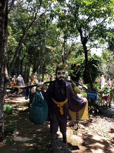 The image depicts a series of statues in an outdoor setting, likely a temple or garden in Nam Ban, Vietnam. The main subject is a large statue of a figure with a beard, wearing a dark robe and a golden sash, holding a staff and a large green bag. Behind this figure, other statues are visible, including one with a smiling face and another that appears to be a Buddha. The scene is set amongst lush green trees, with sunlight filtering through the leaves, casting dappled shadows on the ground.
A blue sign is visible in the background, featuring Vietnamese text and the English words "RESTROOM AREA," indicating this is a public area with facilities. The overall atmosphere is tranquil and spiritual, with the statues and natural surroundings creating a serene environment. The time of day appears to be daytime, with bright sunlight. The statues are detailed and colorful, suggesting they are significant to the location.