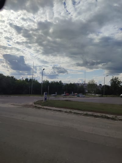 This image depicts an outdoor scene, likely a street or intersection in Barmaky, Ukraine. The foreground shows a paved road with a slight curve to the right, bordered by a grassy area and a concrete curb. In the midground, a dense line of green trees stretches across the scene. Beyond the trees, several modern multi-story buildings are visible in the distance.
Several objects are present. Two tall, slender poles are spaced apart in the middle ground, one significantly taller than the other, possibly telecommunication towers or flagpoles. A lamppost with a single light fixture stands near the center. A blue rectangular sign with a white arrow pointing to the right is situated on a short post. To the left of this sign, there are several large, rectangular containers, some colored red, green, and grey, likely for waste disposal.
The sky is filled with dramatic, heavy gray clouds, suggesting overcast or stormy weather. Patches of blue sky are visible through the clouds in places. The lighting indicates it is daytime, but the cloud cover creates a somewhat subdued atmosphere. The overall impression is that of a slightly somber, cloudy day in an urban or suburban environment. There is no discernible activity or people visible in the image, and no visible text on the objects.