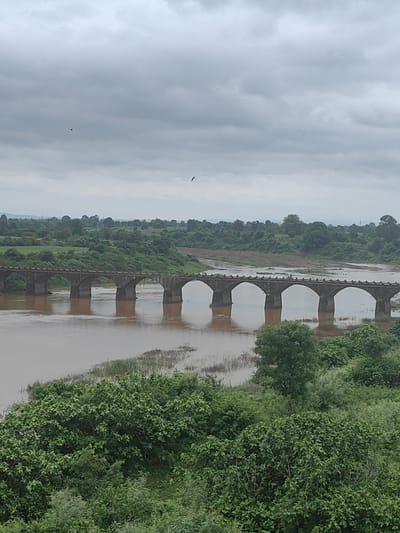 The image shows a cloudy sky over a wide, muddy river and a stone arched bridge. The bridge has multiple arches and is supported by thick pillars. The riverbanks are lined with lush green trees and vegetation. In the distance, there are rolling hills. A small bird is visible in the sky. The scene appears to be natural and rural.
