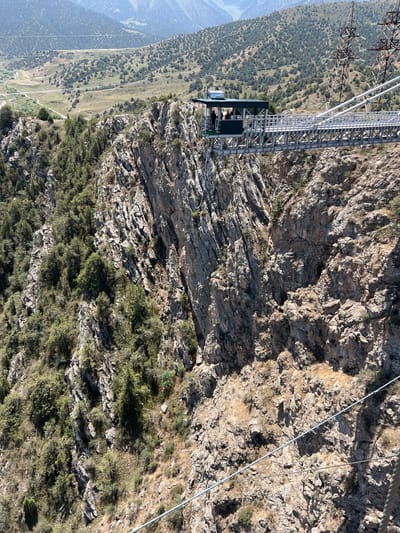 The image shows a bungee jumping platform suspended over a deep canyon in Dardar, Uzbekistan. The platform, a metal structure with a green roof, extends from the rocky cliff face and features a railing along its edge. Several people are visible on the platform, appearing to be participants or staff. The canyon walls are steep and covered with sparse vegetation and large rock formations. In the background, rolling hills covered with trees and some power lines stretch towards distant mountains under a clear blue sky. The overall scene conveys a sense of adventure and dramatic natural scenery.
