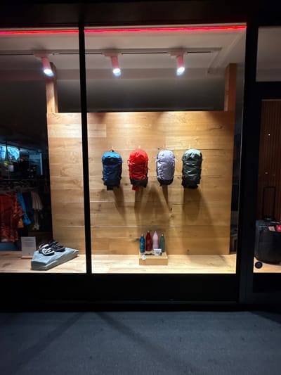The image shows a storefront display at night. Four backpacks are hanging on a wooden wall in the display window. From left to right, the backpacks are dark blue, red, light purple, and dark green. Below the backpacks, there are several colored bottles and a pair of sandals on a grey rock-like display. The storefront is located in the city of Innsbruck, Austria. The street in front of the store is dark, with shadows cast from the display.