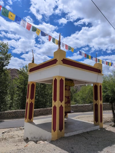 The image captures a prayer wheel structure located in Phyang, India. The structure is painted in yellow and maroon, with a white roof. A large, cylindrical prayer wheel with intricate golden designs is housed within the structure. Prayer flags, a common sight in Buddhist regions, are strung across the sky, adding a splash of color against the bright blue sky dotted with white clouds. Green trees and a stone wall are visible in the background, suggesting a rural or mountainous setting. A paved road runs alongside the prayer wheel structure, indicating its accessibility. The ground is covered with sand and pebbles.
