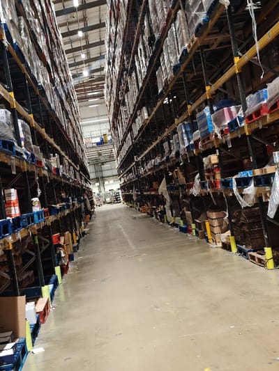 The image depicts an interior view of a commercial warehouse in Dunstable, United Kingdom. A long, concrete aisle extends towards the background, flanked on both sides by multi-level high-bay racking systems. These racks are heavily stocked with palletized goods, predominantly wrapped in clear plastic film, extending upwards towards a high, industrially lit ceiling. The concrete floor of the aisle is clear, showing some minor scuffs and faint markings, with small amounts of debris or packaging materials visible along the base of the racks near yellow floor protection elements. No individuals are present, and no active operations or interactions are observable. Overhead strip lighting illuminates the entire scene.