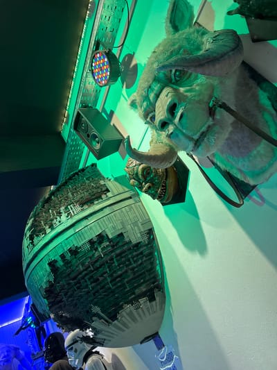 This indoor scene features a collection of Star Wars-themed memorabilia, predominantly illuminated by artificial green and blue lighting. A large, white, furry replica head of a Wampa, complete with horns, hangs prominently from the ceiling, casting a shadow. Below and to the right of the Wampa head, a smaller, greenish creature head (possibly Salacious Crumb) is also displayed.
To the right, a massive, highly detailed replica of the Death Star hangs, showcasing intricate grid patterns and structural components. Partially visible to the far right, two white Stormtrooper helmets are part of the exhibit.
The display includes technical equipment: a black speaker and a multi-colored LED par light are mounted on a metal track running along a dark wall on the left side of the frame. An LED strip light also runs along the bottom edge of this track. The ceiling is a light color, reflecting the green light. A blue light source is visible on the far right, and faintly discernable text "PROHIBIDO" (Spanish for "Forbidden") can be seen on a blue plaque. The word "BLEG" is visible on the metal track. The scene portrays a static display, likely within a themed room, museum, or shop in Villa General Belgrano, Argentina.