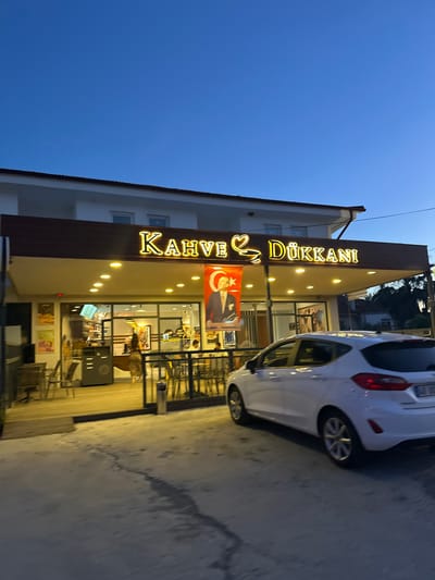 Exterior shot of a "Kahve & Dukkani" coffee shop in Köyceğiz, Turkey. A white car is parked in front of the shop. The facade of the shop has a brown frame. The signage "Kahve & Dukkani" is illuminated with lights. A Turkish flag with a portrait is hanging in the shop window. The sky is dark blue. The shop is open, with tables and chairs visible inside.