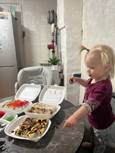 A young child with blonde hair in pigtails is reaching for a piece of sushi on a dark, speckled table. The child is wearing a burgundy shirt with a floral pattern on the sleeves and patterned leggings. Several open white takeout containers filled with different types of sushi are arranged on the table. One container has bright orange roe on top of the rolls, another has what appears to be crispy fried onions drizzled with a dark sauce, and a third contains sushi rolls covered in rice and drizzled with sauce. There is also a small container with green and red condiments, and a lid from a soy sauce dish. The background shows a white tiled wall and exposed pipes with a red valve. A gray high chair is partially visible behind the table. The lighting suggests it is indoors, possibly in a kitchen or dining area. The scene appears casual and domestic. There is no visible text in the image.