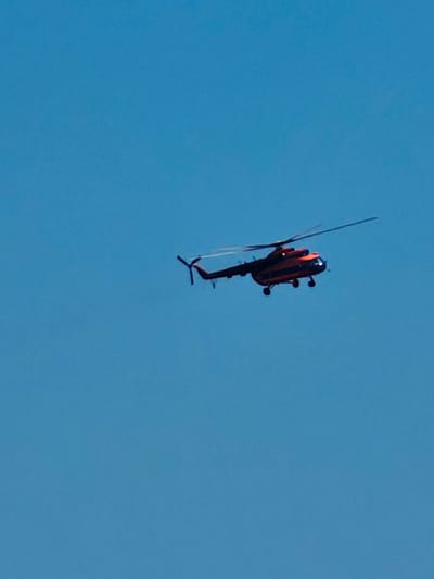 A red and black helicopter is captured in mid-flight against a clear blue sky. The helicopter is angled from the side, showing its rotors and landing gear. There are no other objects or people visible in the frame. The scene is devoid of any specific environmental context that would indicate a location within the city of Yeniseysk, Russia, or any urban setting. The image appears to be an isolated capture of the aircraft in flight.