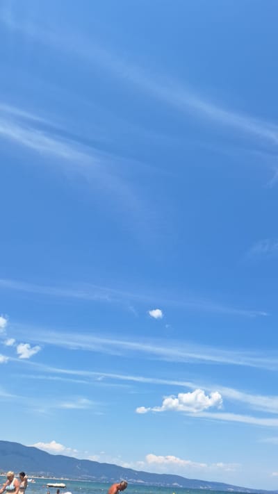The image captures a sunny day at the beach in Stavros, Greece. The sky is a vibrant blue with scattered white clouds, some wispy and elongated, others more puffy. In the background, a range of mountains forms a silhouette against the horizon. The sea is visible, with gentle waves lapping at the shore.
Several people are present on the beach and in the water. In the foreground, a woman with blonde hair is standing in the water, wearing a striped bikini. To her left, another woman in a bikini is also visible. A man with grey hair is bending down in the water, appearing to be engaging with something. Further down the beach, other individuals can be seen, some partially obscured by the water or other beachgoers. There are also some floating objects in the water, possibly inflatable toys or buoys. The overall scene depicts a typical day at a coastal resort.