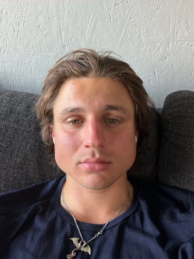 A young man with brown hair and fair skin is seated on a grey couch, facing forward and looking directly at the camera. He is wearing a dark blue t-shirt and a silver chain with a dragon pendant. His left cheek appears swollen. He has earbuds in both ears. The background consists of a textured white wall and the grey couch. The image was taken in Sandavágur, Faroe Islands.