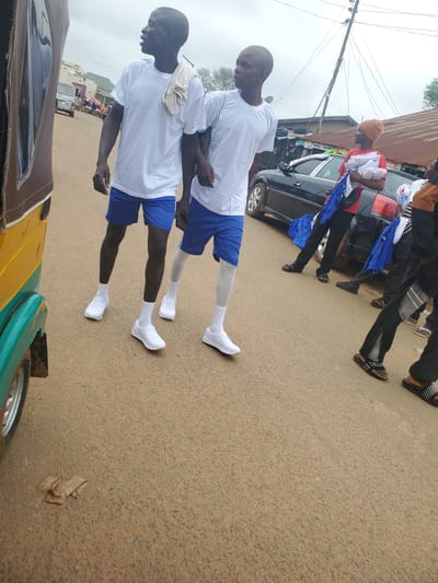 The image depicts a street scene in Ohit, Nigeria, on an overcast day. In the foreground, a portion of a yellow and green auto-rickshaw is visible on the left. The road surface is paved with asphalt and appears to be dusty.

Two young men in matching white t-shirts and blue shorts are the primary subjects. They are both wearing white socks and white athletic shoes. One man, on the left, has a white towel draped over his shoulder. They are both looking to their right, away from the camera.

Behind them, further down the street, several other individuals are present. One person is wearing a red shirt and an orange cap, holding a bundle of white fabric. A black car is parked behind this individual. Other people are seen in the background, some carrying similar bundles of fabric. The environment suggests a commercial or residential area with buildings and utility poles visible. The overall impression is of a bustling street with activity.