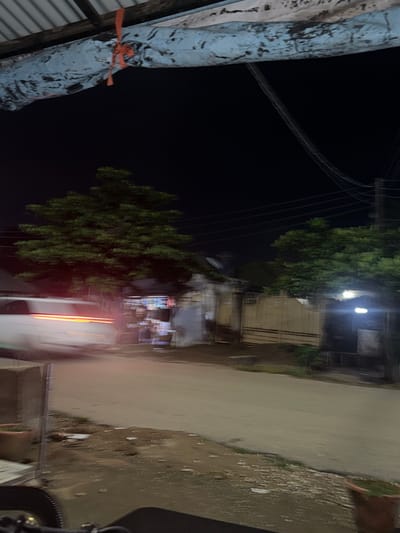 The image depicts a blurry nighttime scene on a paved road in Abuja, Nigeria. A white vehicle, its lights blurred due to motion, is moving from left to right across the frame. To the left of the road, a large green tree stands prominently, its foliage indistinct. Behind the tree, a yellowish gate with horizontal bars is visible, suggesting a residential or commercial property. The sky is dark, characteristic of nighttime. Overhead, power lines stretch across the frame, and a portion of a corrugated metal roof is visible in the upper left corner, partially covered by a blue tarp secured with orange twine. The ground in the foreground appears to be a mixture of dirt and patchy vegetation, with a terracotta pot containing green grass on the far left. The overall impression is of a street scene at night, with the blurriness indicating a long exposure or movement during the capture of the photograph.
