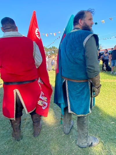 Two men in medieval costumes stand on a grassy field under a clear blue sky. The man on the left wears a bright red tunic with ornate trim and dark trousers, holding a red flag with a white emblem. He wears dark leather boots. The man on the right is dressed in a teal tunic with decorative trim over a green undershirt, and wears a leather belt and gauntlets. He stands with his back to the camera, facing right, and wears tall, grey leather boots. A teal flag is partially visible behind him. In the background, a crowd of people are gathered, some standing and others sitting, with a dog lying down on the grass. Colorful triangular flags are strung across the sky between poles. The scene appears to be an outdoor event, possibly a historical reenactment or festival, and the image location is identified as Llanera, Spain.