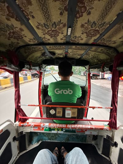The image is a POV shot from inside a tuk-tuk in Khan Boeng Keng Kang, Cambodia. A person wearing a green shirt with a white "Grab" logo is driving. The tuk-tuk has a decorative fabric roof with floral patterns and red curtains on the sides. The road outside is wet, suggesting recent rain, and traffic is visible, including buses and other vehicles. The interior of the tuk-tuk is decorated with various stickers and has a painted mural on the lower section. The legs and feet of the person taking the photo are visible in the foreground, wearing blue jeans and sandals.