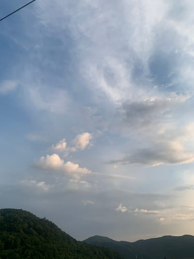 The image displays a dramatic sky with a mix of light and dark clouds, suggesting dynamic weather conditions. The sky is vast and fills most of the frame. On the right side of the image, a densely forested mountain range slopes downwards, its dark green foliage contrasting with the lighter tones of the sky. The lighting indicates it is likely late afternoon or early evening, with the sun's rays creating highlights on some of the clouds. The overall mood is serene and natural, capturing a moment of twilight over a mountainous landscape. The scene appears to be a natural setting, possibly a remote area given the lack of visible man-made structures besides a thin black line that might be a cable or wire in the lower left corner. No people or specific activities are depicted. There is no visible text in the image.