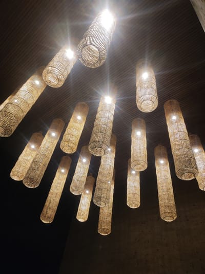 This image captures an interior space in Mas, Indonesia, featuring a cluster of approximately 19 cylindrical pendant lights suspended from a dark ceiling. Each light fixture is constructed from woven natural fibers, likely rattan or bamboo, enclosing an illuminated bulb that emits a warm, yellowish-white light. Several light sources exhibit prominent lens flares. The ceiling above the lights is dark with a subtle horizontal texture, possibly slats or grooves. A dark, rough-textured wall, consistent with concrete or stone, is partially visible on the right side of the frame. No human figures or discernible actions are present.