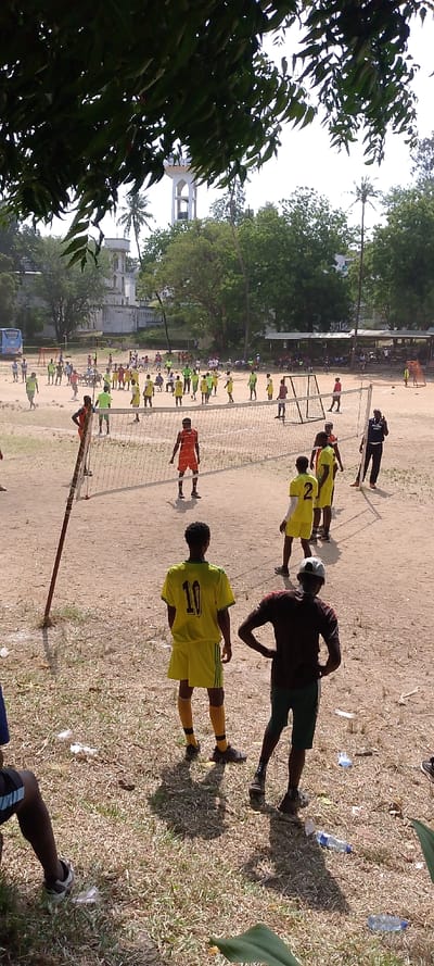 The image depicts an outdoor sports field in Tudor ward, Kenya, under clear daylight. In the foreground, partially obscured by dark green foliage at the top left, a dusty, dry earth playing surface is visible, sparsely covered with dry grass and scattered litter, including plastic bottles. Three individuals are prominently positioned in the lower-right foreground: one young male from the waist down, another young male wearing a yellow jersey with the number "10" on the back and matching shorts and socks, facing away from the camera, and a third young male in a dark t-shirt and dark shorts, hands on hips, also facing away.
Mid-ground, a volleyball net is stretched across the field, supported by poles. On either side of the net, groups of young males in sports attire (primarily yellow and orange jerseys) are engaged in what appears to be a volleyball game or practice. Further back on the field, numerous other individuals, predominantly young males, are visible. Many are wearing bright green or yellow jerseys, participating in what appear to be football (soccer) activities, with at least one soccer goal frame visible. A blue bus is parked on the far left side of the field. To the left of the field, a large, light-colored building with multiple stories, crenellated walls, and towers stands amidst trees.
The background features a collection of green trees and structures. Above the tree line, a tall, white water tower with an open-frame top is visible on the right. Another, smaller, white architectural structure with a dome or distinct top feature is partially visible on the left, behind trees. The sky is bright and clear.