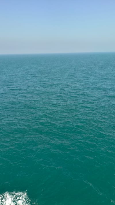 This aerial photograph captures a serene ocean scene. The image is divided vertically, with the left side showing a soft, hazy blue sky that gradually transitions into the ocean. The right side of the image is dominated by the deep, turquoise water, which exhibits a textured surface with gentle ripples. The transition between the sky and the water is blurred, suggesting a hazy or misty atmosphere, possibly indicating early morning or late evening light.
In the lower right corner, a small patch of white, foamy water is visible, suggesting movement, perhaps from a boat or a wave. There is no discernible human activity or specific objects in the frame, besides the implication of a moving watercraft. The overall mood is tranquil and expansive, emphasizing the vastness of the ocean and the sky. The lighting is diffused, and the colors are muted, contributing to a sense of peacefulness. No text is visible in the image.