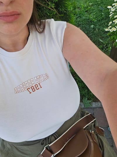 In Petropavlivska Borshchahivka, Ukraine, a close-up, overhead shot captures a woman wearing a white t-shirt with a distressed "SANTA BARBARA 1981" graphic in brown lettering. She also wears olive-green pants and a brown crossbody bag with a white and brown strap. The background features lush green grass and small white flowers, suggesting an outdoor, possibly park or garden setting. The lighting is natural and the overall mood is casual.