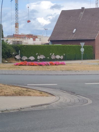 The image depicts a street scene in Vreden, Germany, with a construction site visible in the background. A large yellow crane with a red load hangs in the sky. In the middle ground, a row of pink and orange flowers are planted in front of a tall green hedge. Several abstract metal sculptures resembling flamingos are placed among the flowers. A brick house with a brown tiled roof and a dormer window stands to the right of the hedge. A street with a paved pedestrian path curves through the foreground, bordered by dry grass and a concrete curb. The sky is cloudy.