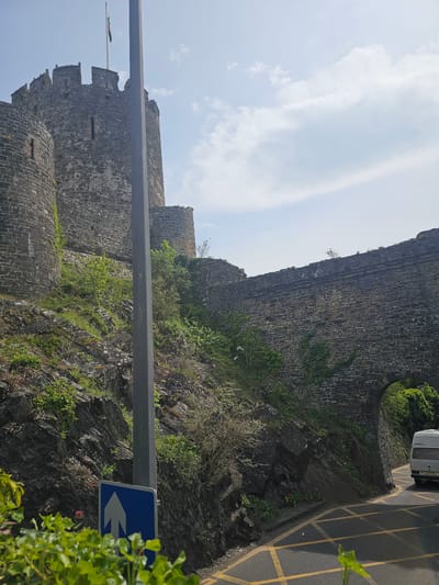 The image depicts a section of Conwy Castle in Llandudno Junction, United Kingdom, framed by a street sign and a roadside wall. The castle, built from stone, features crenellations and window openings. Vegetation grows on the castle walls and surrounding rock face. A road with double yellow lines and a parked white vehicle is visible at the top right. The sky is partly cloudy.