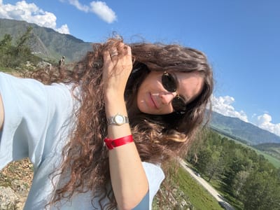A selfie taken outdoors features a woman with long, curly brown hair and sunglasses. She is wearing a light blue t-shirt and a watch on her left wrist, along with a red wristband. Her hand is in her hair. The background shows a mountainous landscape with green slopes and a road winding through it, suggesting the location is near Bichiktu-Boom, Russia. The sky is blue with scattered clouds.