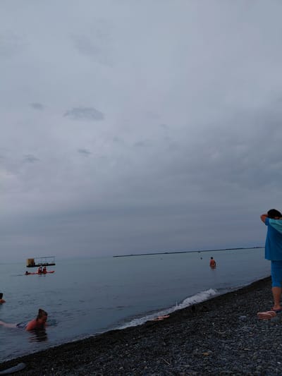 The image depicts a cloudy, overcast day at a pebble beach in Ақши, Kazakhstan. In the foreground, a person in a blue t-shirt and blue leggings stands with their back to the camera, their arms raised to their head. Scattered footwear, including sandals and shoes, lies on the pebble shore. To the left, a woman with her hair in a bun is partially submerged in the water, appearing to be swimming or wading. Further out in the water, a person stands in shallow water, and another is seen from the back in the water. In the mid-ground, a raft-like structure with a canopy is visible, with at least two individuals on it. Two orange kayaks are also present in the water near the raft, with at least two people in them, one appearing to be wearing a life vest. The sea stretches out to the horizon under a dense, gray sky. In the far distance, a thin, dark line suggests a distant shore or structure. The overall scene conveys a subdued, possibly cool, atmosphere with limited activity.