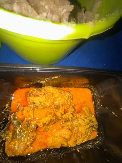 The scene depicts two distinct food items presented on a blue surface in Ilishan, Nigeria. In the upper portion, a light green bowl, partially cropped, contains a quantity of light-colored, granular fufu (likely pounded yam). Below it, a black rectangular plastic takeaway container holds a reddish-orange stew with several pieces of protein, possibly meat or fish, submerged within the sauce. The stew exhibits an oily sheen and a textured consistency. No individuals are visible. The items are arranged consistent with a meal ready for consumption.
