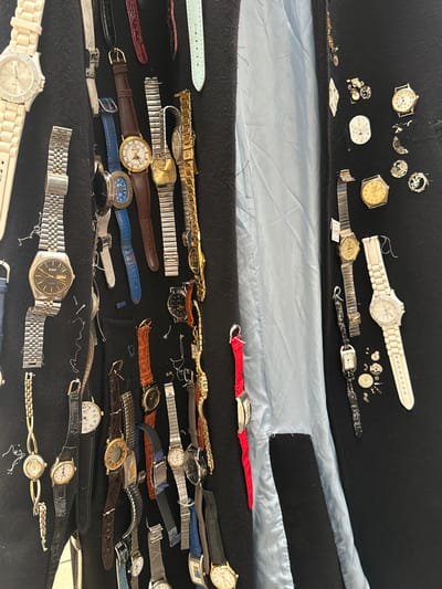 The image shows a collection of watches, watch parts, and watch straps arranged on a black fabric surface. Some are intact watches with bands, while others are disassembled, with parts scattered around. A blue-grey material is visible beneath the black fabric in the upper part of the image. The scene appears to be a disorganized collection of timekeeping devices and related accessories. There is no information available in the image to suggest a location in the city of Montreuil, France.