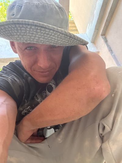 A man wearing a light-colored bucket hat and a dark t-shirt is crouched down, looking towards the camera. His face is partially obscured by the brim of his hat. He has light-colored eyes and appears to be outdoors, with some vegetation visible in the background. The setting appears to be an unfinished concrete structure, possibly under construction. The man's left arm is bent, and his right arm is resting on his lap. The image was taken at an angle looking up at the man.